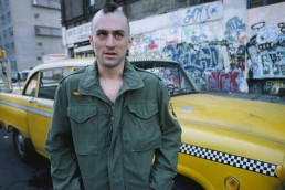Robert De Niro as Travis Bickle standing beside a yellow taxi in Taxi Driver (1976)